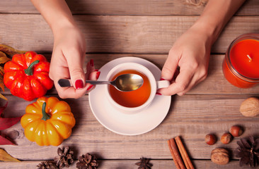 Woman's hand holding spoon and cup of tea. Autumn leaves, candle, pumpkin on the wooden background.