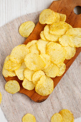 Salted wavy potato chips on a rustic wooden board on a white wooden surface, top view. Overhead,...