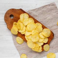 Salted wavy potato chips on a rustic wooden board on a white wooden background, overhead view. Top...
