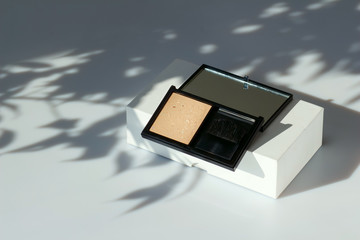 Fototapeta premium Face powder in a black case. Fashion cosmetics on a white background with a shadow of leaves. Open eyeshadow, compact foundation. Concealer face powder, blush. Cosmetics with natural ingredients.