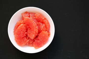 Red grapefruit slices in a white bowl on a black surface, top view. Flat lay, overhead, from above. Space for text.