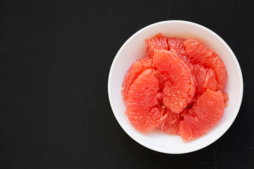 Red grapefruit slices in a white bowl on a black background, top view. Flat lay, overhead, from above. Copy space.