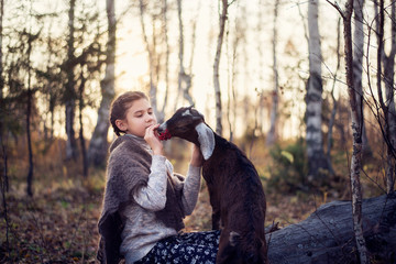 A cute girl sits in a forest on a fallen tree and feeds a goat.