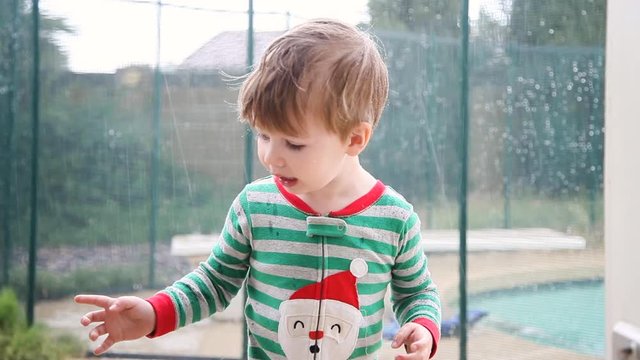 Little boy dressed in Christmas pajamas stands outside in the rain. He is not sure what to make of the sprinkles coming down from the sky and shakes his arms and hands. He winces and makes expressions