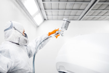 Auto mechanic worker painting a white car with spray gun in a paint chamber during repair work.