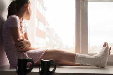 A teenage girl with a broken leg in a cast is sitting on a windowsill with crutches.