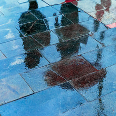 Blurry reflection shadows silhouettes of a woman under umbrella and a man walking in the rain in the city sidewalk, autumn rainy evening