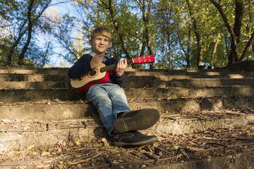 A boy in a blue jacket and jeans plays the guitar on the old steps in an abandoned park.