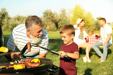 Grandfather with little boy cooking food on barbecue grill and their family in park