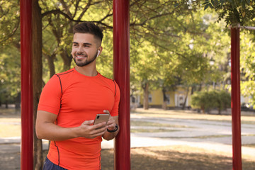 Young man with wireless headphones and mobile device listening to music on sports ground. Space for text