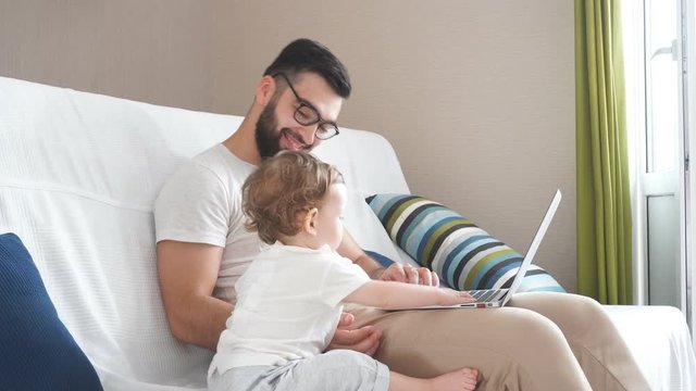 Bearded clever handsome man in glasses working on the laptop, while his baby helping him in the bedroom, job, profession, free time