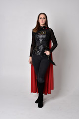 full length portrait of a pretty brunette woman wearing black leather fantasy costume with long red superhero cape. standing pose on a studio background.