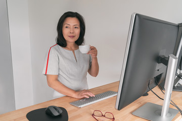 Mature Asian business woman working on standing desk at home office drinking coffee cup taking a...