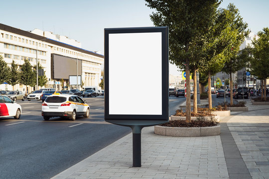 Vertical Commercial Banner Stand With Blank Digital Screen To Advertise Products And Services. Billboard For Ads In City Center. Board For Announcements Near Highway With Busy Traffic, Buildings.