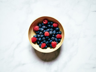 Blueberries and Raspberries in a wooden bowl on a white marble background viewed from above