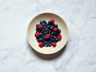 Blueberries and Raspberries in a cream dish on a white marble background viewed from above
