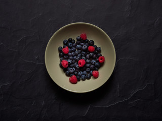 Blueberries and Raspberries in a cream dish on a dark stone background viewed from above