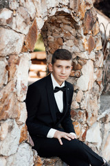 Teen guy in stylish black suit with bow tie. Summer holiday and vacation concept. Happy stylish school boy posing in a stone ring. Portrait of handsome young male student outdoors. 