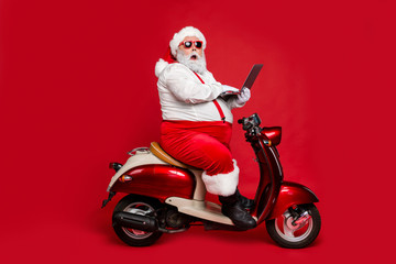Obraz na płótnie Canvas Profile side view of his he nice fat wondered funky Santa sitting on moped holding in hand laptop reading wish dream list winter fairy isolated on bright vivid shine vibrant red color background