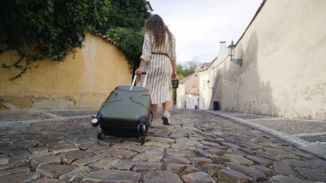 Following shot female going along narrow street with travel luggage in sunny day. Back view young girl rolling suitcase on the cobblestone pavement in downtown of European tourists town.