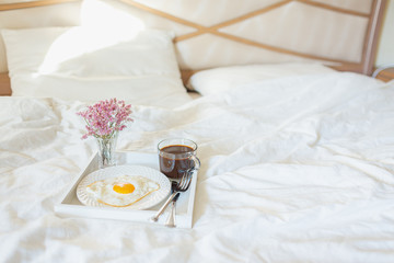 White tray with breakfast on a bed in a hotel room. Fried egg, cup of coffee and flowers in white sheets in light bedroom. Copyspace.