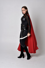 full length portrait of a pretty brunette woman wearing black leather fantasy costume with long red superhero cape. standing pose on a studio background.