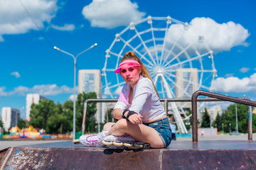 Portrait of an emotional girl in a pink cap visor wearing protective gloves and rollerblades sitting on the background of ferris wheel.
