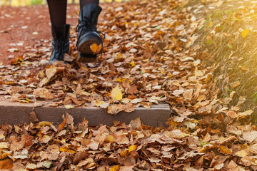A girl in black boots and a red coat kicks yellow and red foliage strolling in the park alone on a clear autumn day during a fall. Freshness, nature and fun.