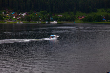 a boat floats to moor to the shore on which there are green trees and other ships in Altai Mountains. Leisure and outdoor travel without people.
