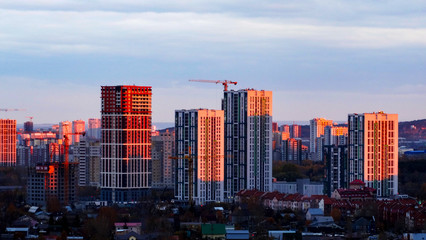 two building cranes at the background of a multi-storey building under construction with some small workers building in front of them against sunset sky. New houses will wait for families