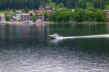 a boat floats to moor to the shore on which there are green trees and other ships in Altai Mountains. Leisure and outdoor travel without people.