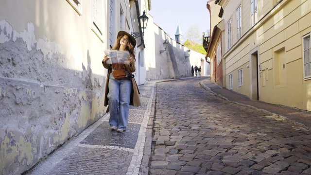 Young woman walking at downtown of Prague. Lady walking down on narrow street in sunny day. Girl checking the map and enjoying old city centre while going on cobblestone pathway between houses