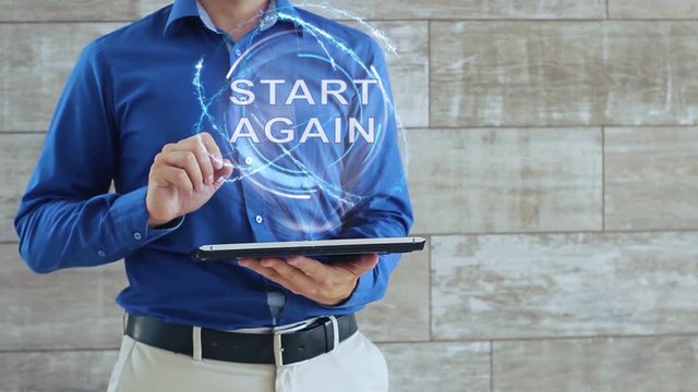 Man activates a conceptual hologram with text Start Again. The guy in the blue shirt and light trousers with a holographic screen gadget on the background of the wall