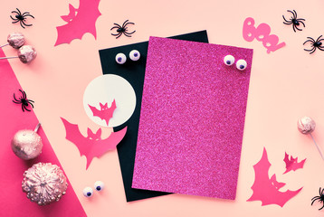Creative paper craft Halloween flat lay in pink, magenta and black. Top view with cipy-space on stack of cards, bats, chocolate eyes, pumpkins and and text "Boo" on split paper background.