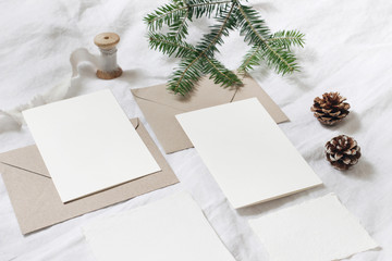Christmas table, festive styled composition. Christmas greeting cards, envelopes mock-ups. Silk ribbon, pine cones and fir tree branch on white linen background. Top view. Festive craft, stationery.