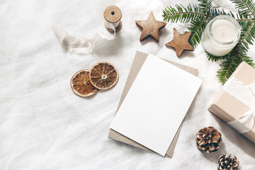Christmas blank greeting card mock-up scene. Festive winter wedding composition. Craft envelope, pine cone, gift box, orange fruit slices and fir tree branch on white table, linen background. Flat lay