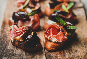 Party or catering food concept. Crostini with prosciutto, goat cheese and grilled figs on wooden board, selective focus, close-up, horizontal composition - obrazy, fototapety, plakaty