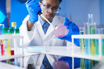 Young African-American man holding petri dish to camera while working on medical research in...