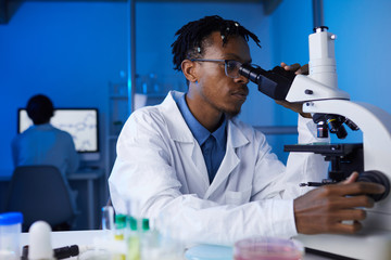 Portrait of young African-American man looking in microscope while working on medical research in...