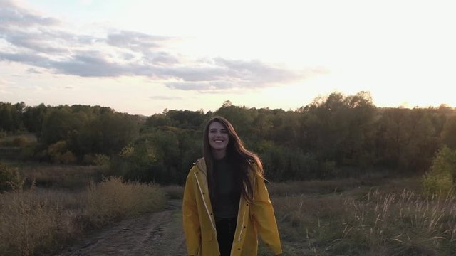 girl in yellow raincoat and rubber boots walks backwards down road and smiles. woman straightens her hair and laughs. Sunlight and glare in camera. evening lighting. Autumn landscape at sunset.
