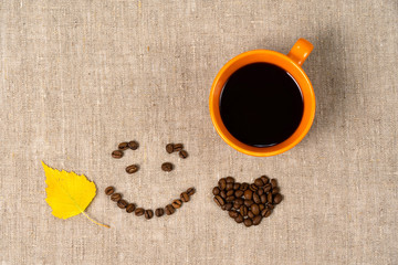 A mug of hot black coffee, coffee beans in the shape of a smile and a heart on a fabric background. Dry fallen yellow leaves, autumn theme