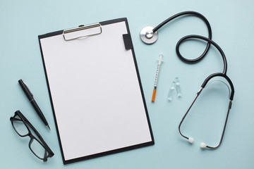Blank white paper on clipboard near stethoscope; injection; pen and spectacles over blue desk