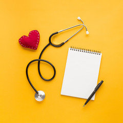 Blank spiral notepad with pen; stitched heart shape; stethoscope above bright yellow backdrop