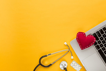 Stitched heart shape on laptop with stethoscope; blister packed tablet on yellow background