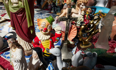 Vintage colorful clown figure raising his fist among other religious porcelain and bronze statuettes standing on the ground at an antiques flea market in Lisbon.