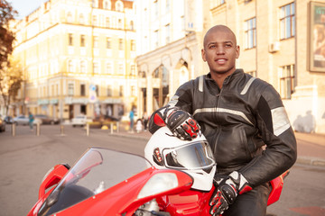 Fototapeta na wymiar Attractive young African man looking around in the city, sitting on his motorbike. Handsome male biker travelling Europe on his motorcycle