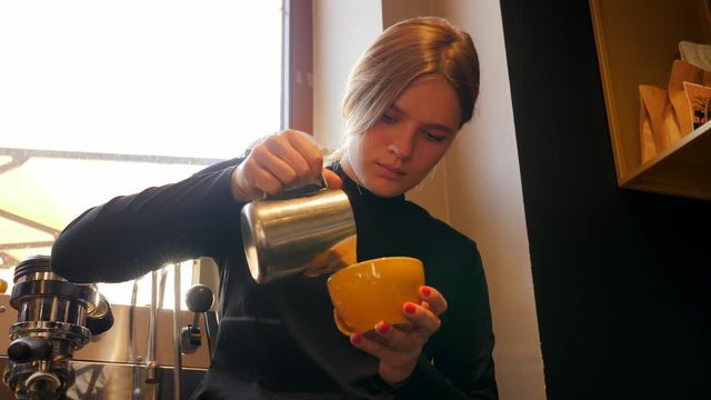 Cute young barista girl in cafe makes coffee cappuccino latte art drawing with milk in the morning