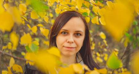 Portrait of a beautiful smiling woman is standing in the autumn forest between the branches of trees with yellow foliage.