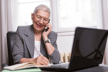 Happy senior old business lady writing down orders, talking on her cell phone