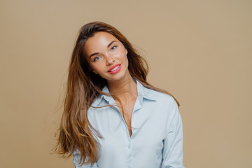 Portrait of pleasant looking cheerful female has long hair, tilts head and looks with smile at camera, wears blue shirt, uses cosmetics for makeup, poses against brown background, blank space aside